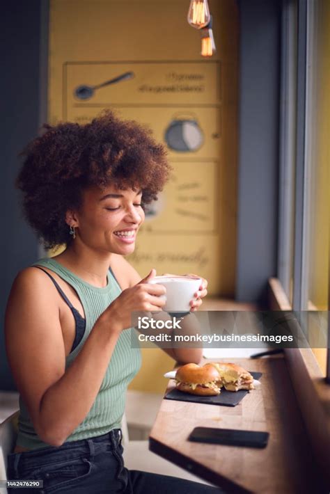 Female Customer In Coffee Shop Window Eating Bagel Sandwich For Lunch